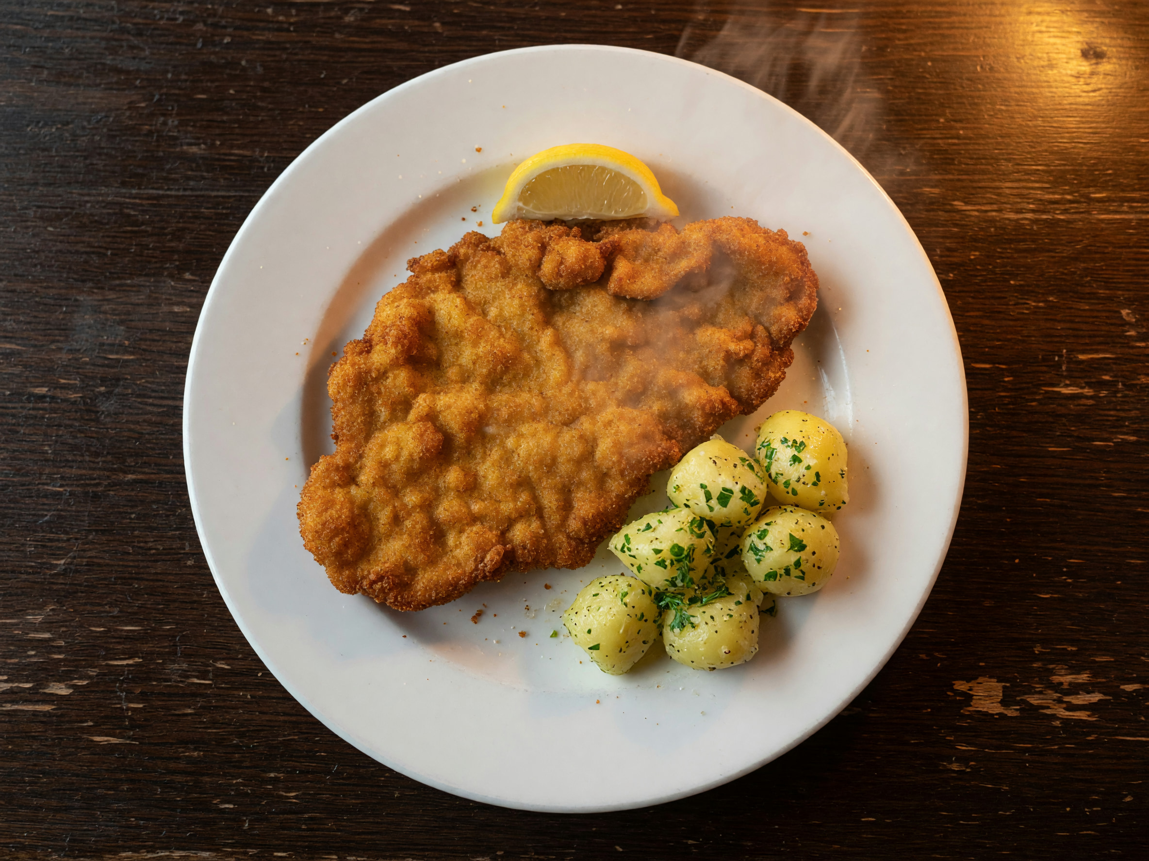 Goldbraun gebackenes Schnitzel mit Zitronenspalte und Petersilkartoffeln auf weißem Teller